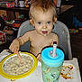 toddler, baby, high_chair, food, spoon, messy, blue_eyes, sippy_cup, indoor, table, plate, child, face, feeding, clutter, toy, drink, expression, person, meal