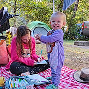 Maia is registered to the contest to win money with this photo: child, toddler, outdoor, camping, tent, markers, drawing, blanket, smile, nature, trees, toy, hat, clothing, person, grass, daylight, parked_vehicle, recreation, leisure