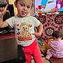 child, toddler, baby, indoor, table, toy, television, carpet, clothing, person, play, furniture, pattern, red_pants, pink_socks, curly_hair, gesture, tongue_out, smile, living_room