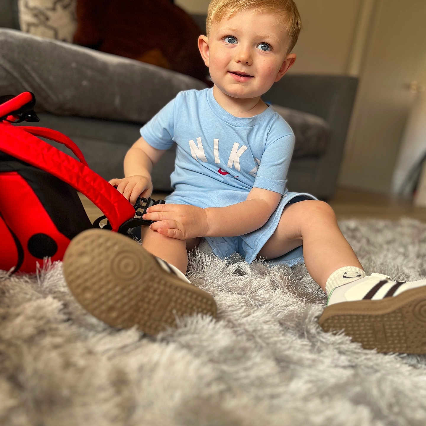 Hugo joined the competition — help win amazing prizes! blue_eyes, carpet, casual, child, curly_hair, cute, home, indoor, light_blue_clothing, living_room, nike, person, playful, red_backpack, rug, sitting, smiling, sneakers, toddler, young_child