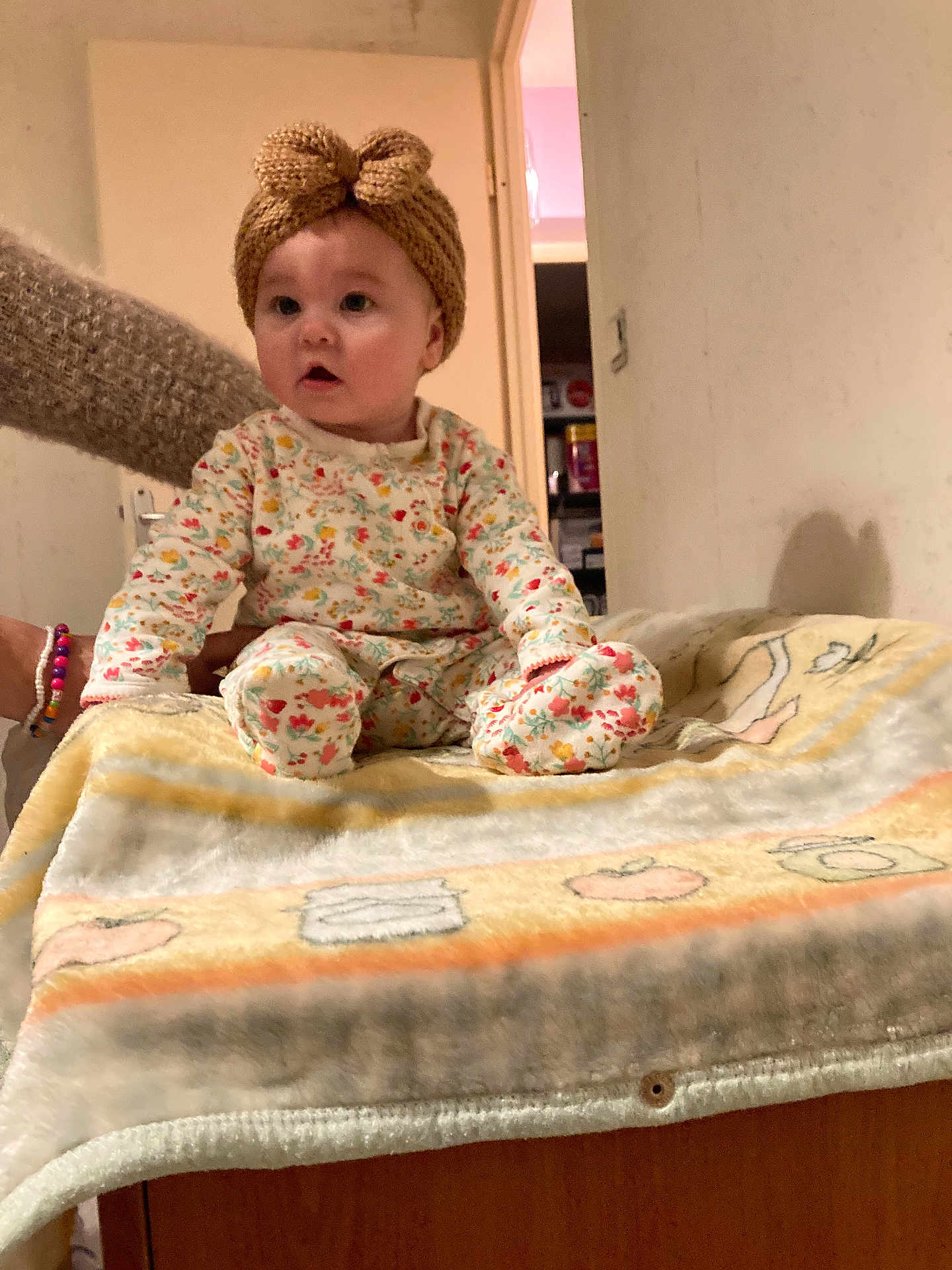 Katalina participe au concours pour gagner de l'argent avec cette photo : baby, child, knitted_headband, bow, blanket, wooden_surface, indoor, person, sitting, long_sleeve_pajamas, patterned_clothing, hand, arm, doorway, pantry, storage, curious, wide_eyes, soft_texture, warm_colors