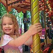 Amarra is registered to the contest to win money with this photo: amusement_park, blonde_hair, butterfly_pattern, carousel, child, colorful, curly_hair, daylight, fence, fun, golden_pole, happy, holding, lights, outdoor, person, pink_shirt, rides, smiling, trees