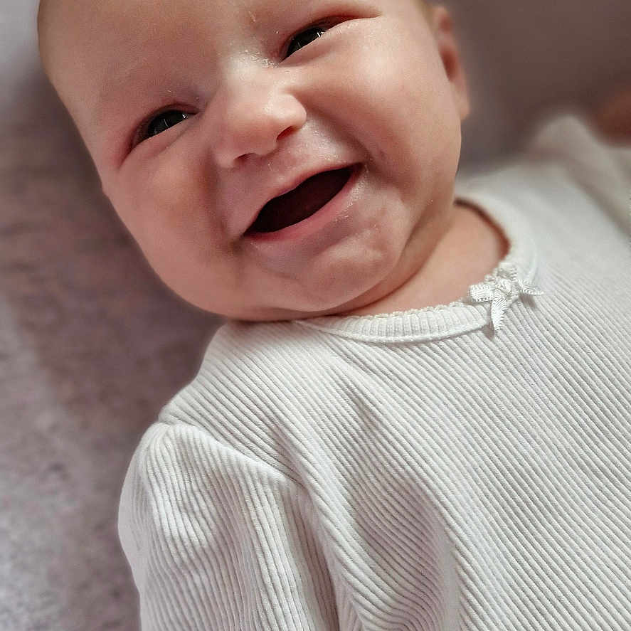 Adah-Jayne is registered to the contest to win money with this photo: baby, child, closeup, cute, expression, eyes, face, happy, head, indoor, infant, joyful, lying_down, mouth, portrait, ribbed_clothing, skin, smiling, soft_background, white_shirt