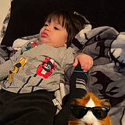 Zayden is registered to the contest to win money with this photo: child, bed, blanket, shirt, car_print, guinea_pig, sunglasses, thumbs_up, digital_sticker, cute, indoors, relaxed, black_pillow, toy, comfort, cozy, playful, humor, animal, person