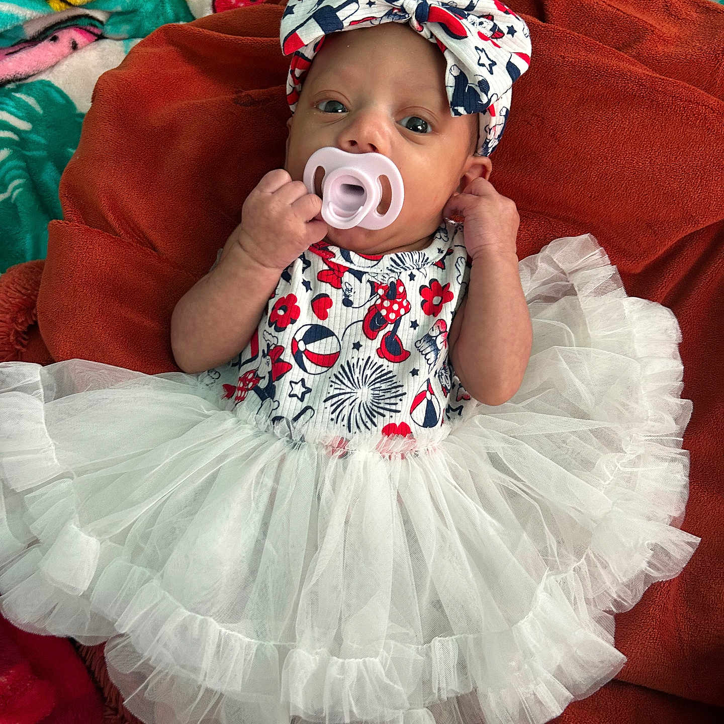 Leeana joined the competition — help win amazing prizes! baby, blanket, blue, bow, colorful, cute, dress, fabric, headband, indoor, infant, lying_down, pacifier, patterned_clothing, person, red, small_hands, soft_texture, tutu, white