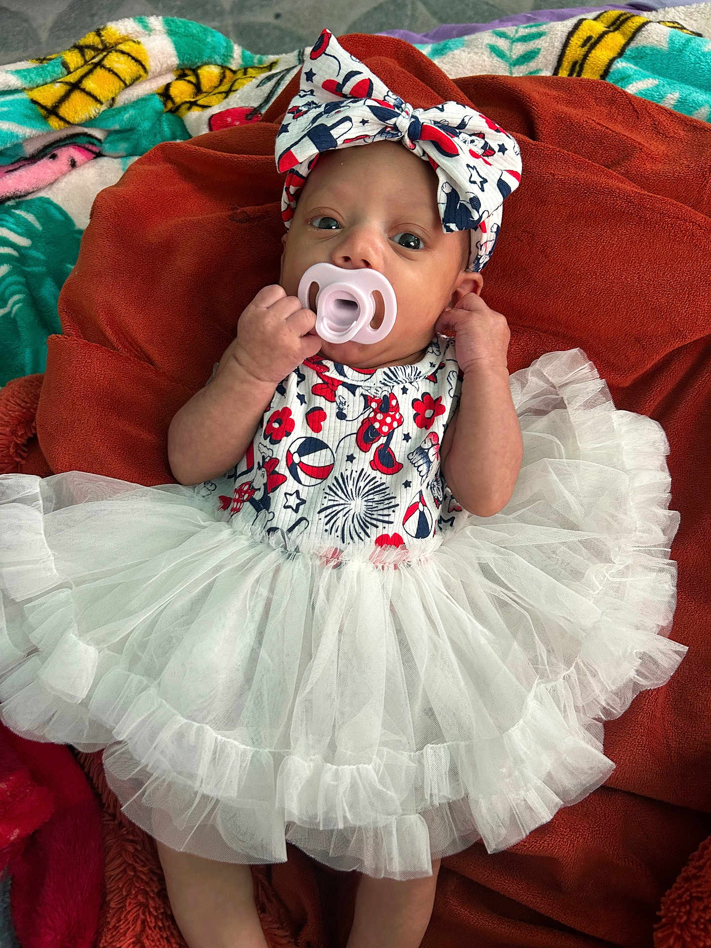 Leeana joined the competition — help win amazing prizes! baby, infant, pacifier, headband, bow, tutu, dress, blanket, patterned_clothing, red, white, blue, lying_down, cute, small_hands, indoor, fabric, colorful, soft_texture, person