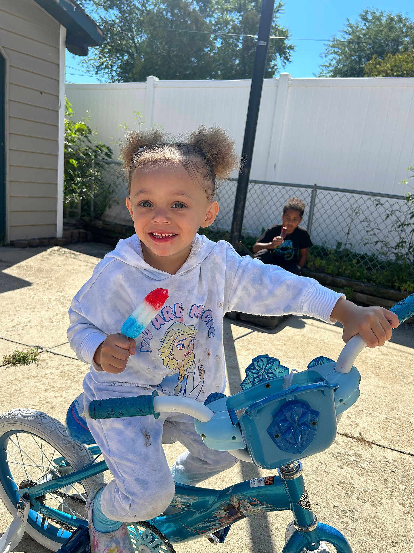 Leigha is registered to the contest to win money with this photo: child, bicycle, popsicle, outdoor, sunny, happy, smiling, curly_hair, hoodie, pants, frozen_theme, blue_bicycle, backyard, fence, person, play, summer, food, fun, toy