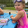 child, toddler, girl, portrait, outdoor, park, picnic, picnic_table, swimsuit, pink_clothing, mermaid_costume, goggles, wet_hair, blue_table, snacks, playground, fence, trees, summer, sitting