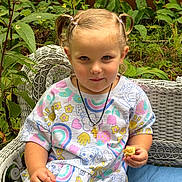 Maggie is registered to the contest to win money with this photo: child, toddler, girl, portrait, smiling, seated, outdoor, garden, wicker_chair, colorful_clothing, pigtails, blonde_hair, necklace, snack, food, plants, leaves, nature, photograph, happy