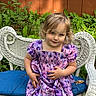 child, girl, toddler, portrait, sitting, smiling, face, blonde_hair, purple_dress, floral_dress, wicker_chair, blue_cushion, garden, plants, outdoor, wooden_fence, 自然, photography, looking_at_camera, colorful