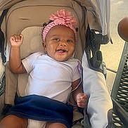 Essence is registered to the contest to win money with this photo: baby, smiling, stroller, headband, flower, white_shirt, bottle, infant, child, happy, raising_hand, footwear, outdoor, seat, fabric, person, cute, baby_girl, toy, comfort