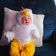 Kendra is registered to the contest to win money with this photo: baby, costume, chicken_costume, yellow_pants, white_fluffy, couch, pillow, indoor, child, face, smiling, seated, cute, soft_texture, plush, person, cozy, home, portrait, funny