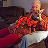 blanket, brown_dog, casual_clothing, comfort, couch, dog, elderly_person, flower_decoration, framed_pictures, glasses, halloween_theme, home_interior, indoor, living_room, orange_outfit, pet_clothing, relaxing, seated, small_dog, striped_shirt
