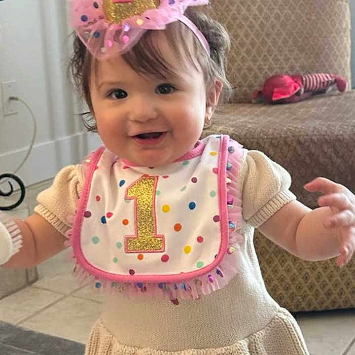 Raylene joined the competition — help win amazing prizes! baby, child, birthday, party_hat, bib, cream_dress, indoor, standing, smiling, happy, floor, armchair, sock, pom_pom, knitted_clothing, celebration, cute, one_year_old, toddler, person