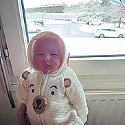 Liam participe au concours pour gagner de l'argent avec cette photo : baby, newborn, infant, onesie, bear_hood, hood, window, snow, cars, parking_lot, adult_hand, hand, radiator, indoors, portrait, sleepy, face, cozy, soft_texture, child