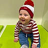 baby, casual, child, clothing, cute, face, green_surface, hand, happy, hat, indoor, infant, jeans, pom_pom, portrait, red_and_white, sitting, smile, socks, striped_shirt