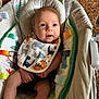 baby, infant, bib, dinosaur_pattern, baby_seat, cushion, cute, child, indoors, carpet, expression, small_hands, small_feet, toy, fabric, comfort, colorful, pattern, curious, portrait
