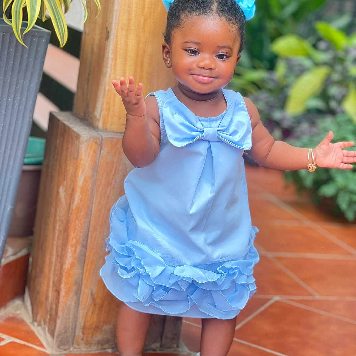 Jazlynn is registered to the contest to win money with this photo: child, toddler, girl, portrait, smiling, standing, blue_dress, hair_bows, blue_bows, sandals, earrings, bracelet, outdoor, patio, tiled_floor, wooden_post, potted_plant, greenery, fashion, full_body