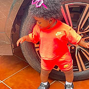 Jazlynn is registered to the contest to win money with this photo: child, toddler, curly_hair, sunglasses, orange_outfit, sportswear, nike, shorts, shirt, shoes, clogs, socks, standing, car, wheel, tire, vehicle, outdoor, tile_floor, portrait