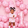 backdrop, balloons, birthday, bows, braids, carpet, celebration, child, floral_pattern, indoor, number_one, party, photography, pink, portrait, smiling, socks, standing, sweater, toddler