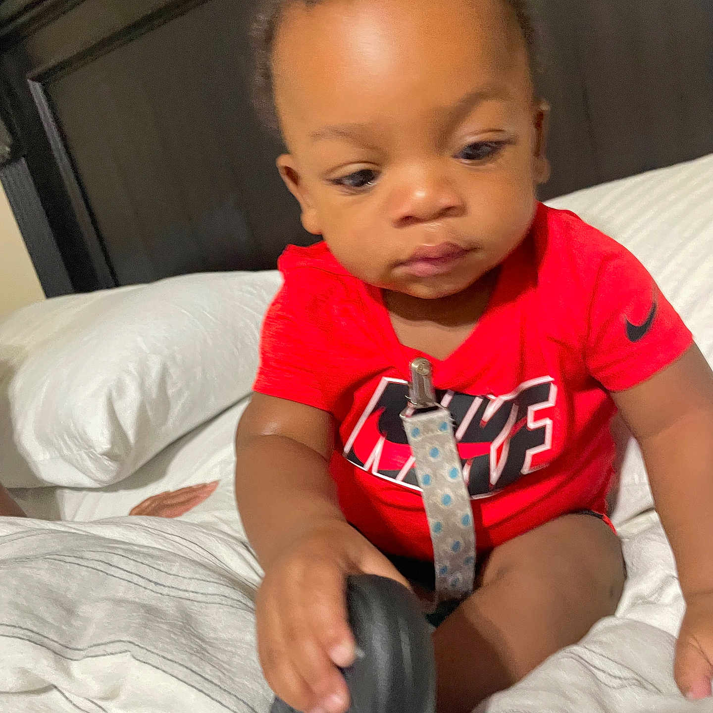 Emmanuel is registered to the contest to win money with this photo: baby, child, infant, portrait, sitting, bed, bedroom, pillow, blanket, red_shirt, black_object, toy, face, eyes, short_hair, serious_expression, indoors, home, photography, nike_logo