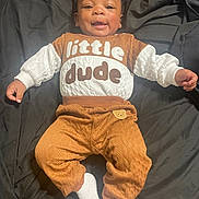 Emmanuel is registered to the contest to win money with this photo: baby, infant, child, smiling, portrait, lying_down, bed, bedding, dark_background, brown_clothing, white_sleeves, quilted_fabric, pants, socks, fashion, casual_clothing, happy, indoors, human, photograph