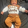 baby, infant, child, smiling, portrait, lying_down, bed, bedding, dark_background, brown_clothing, white_sleeves, quilted_fabric, pants, socks, fashion, casual_clothing, happy, indoors, human, photograph