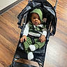 Emmanuel is registered to the contest to win money with this photo: baby, child, infant, stroller, green_outfit, hood, sitting, indoors, wooden_floor, portrait, face, eyes, hands, socks, baby_clothing, harness, black_stroller, floor, photography, cute