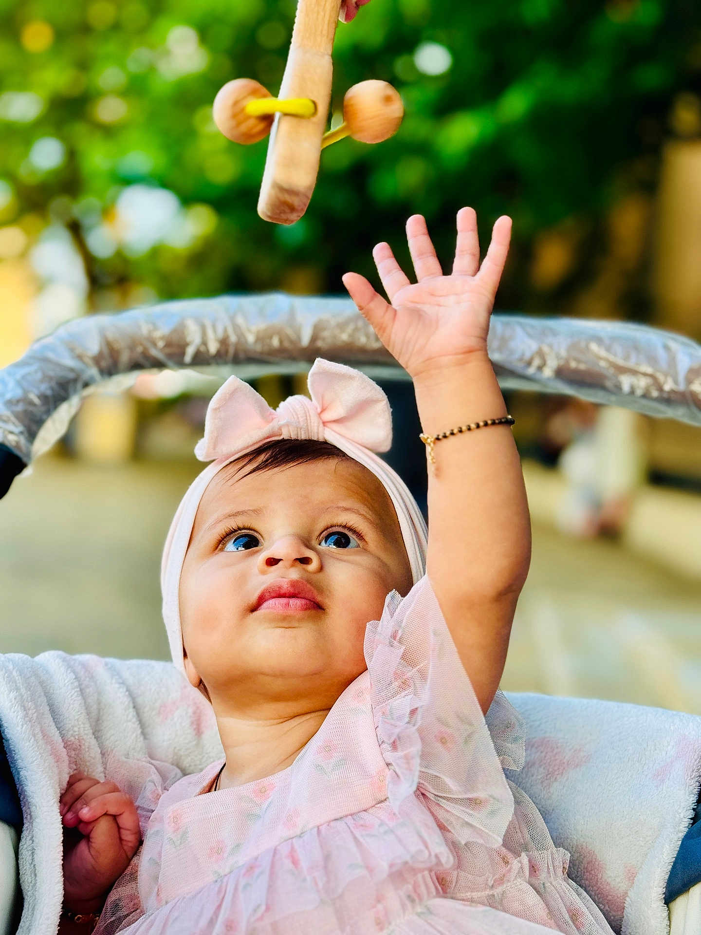 Pahal joined the competition — help win amazing prizes! baby, child, infant, pink_dress, headband, bow, hand, bracelet, stroller, toy, wooden_toy, outdoor, blurred_background, curious, looking_up, face, portrait, candid, soft_light, cute