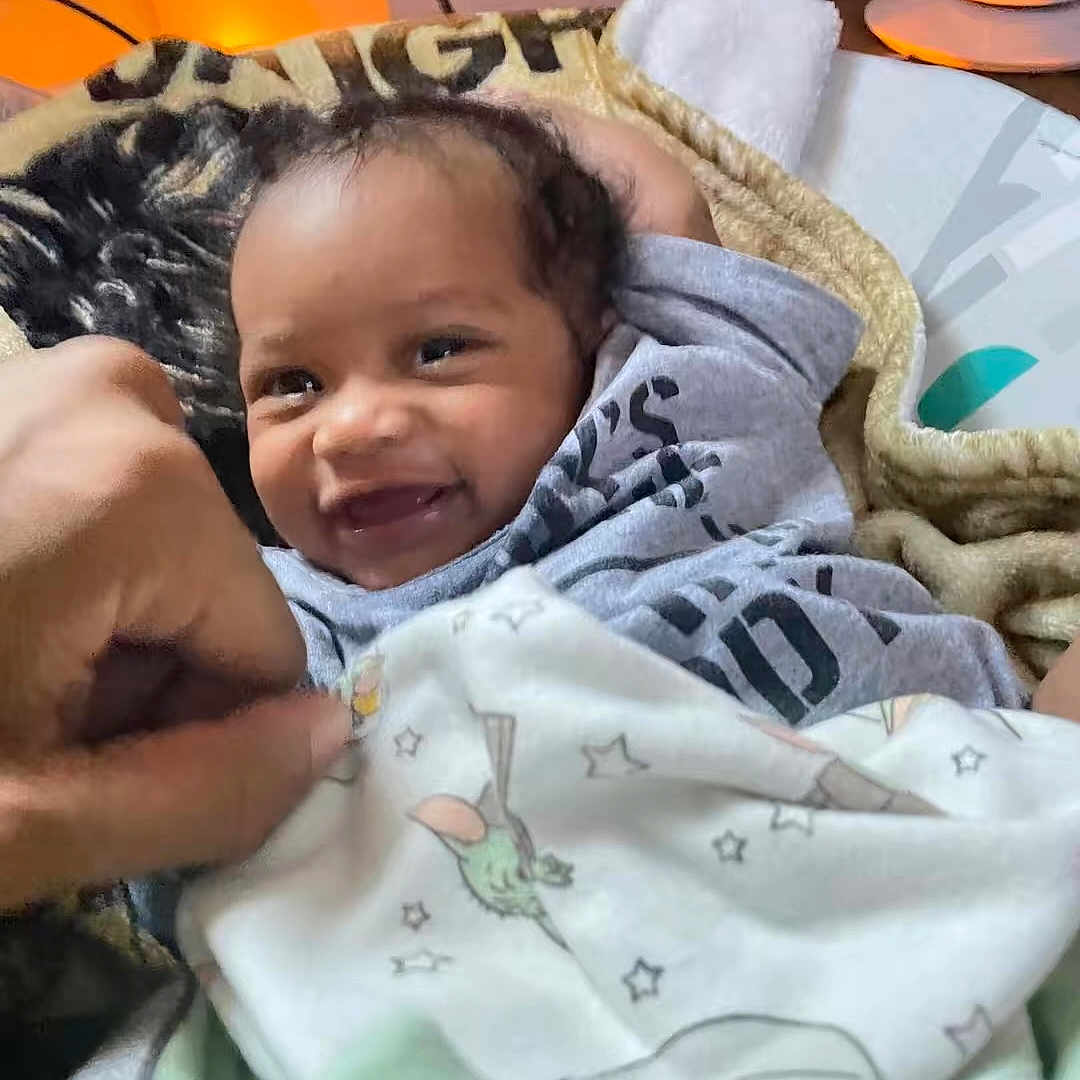 Nyzaiah is registered to the contest to win money with this photo: baby, blanket, clothing, comfort, cozy, cute, expression, face, furniture, hand, happy, indoor, infant, person, playful, portrait, smiling, soft, toddler, warm_light