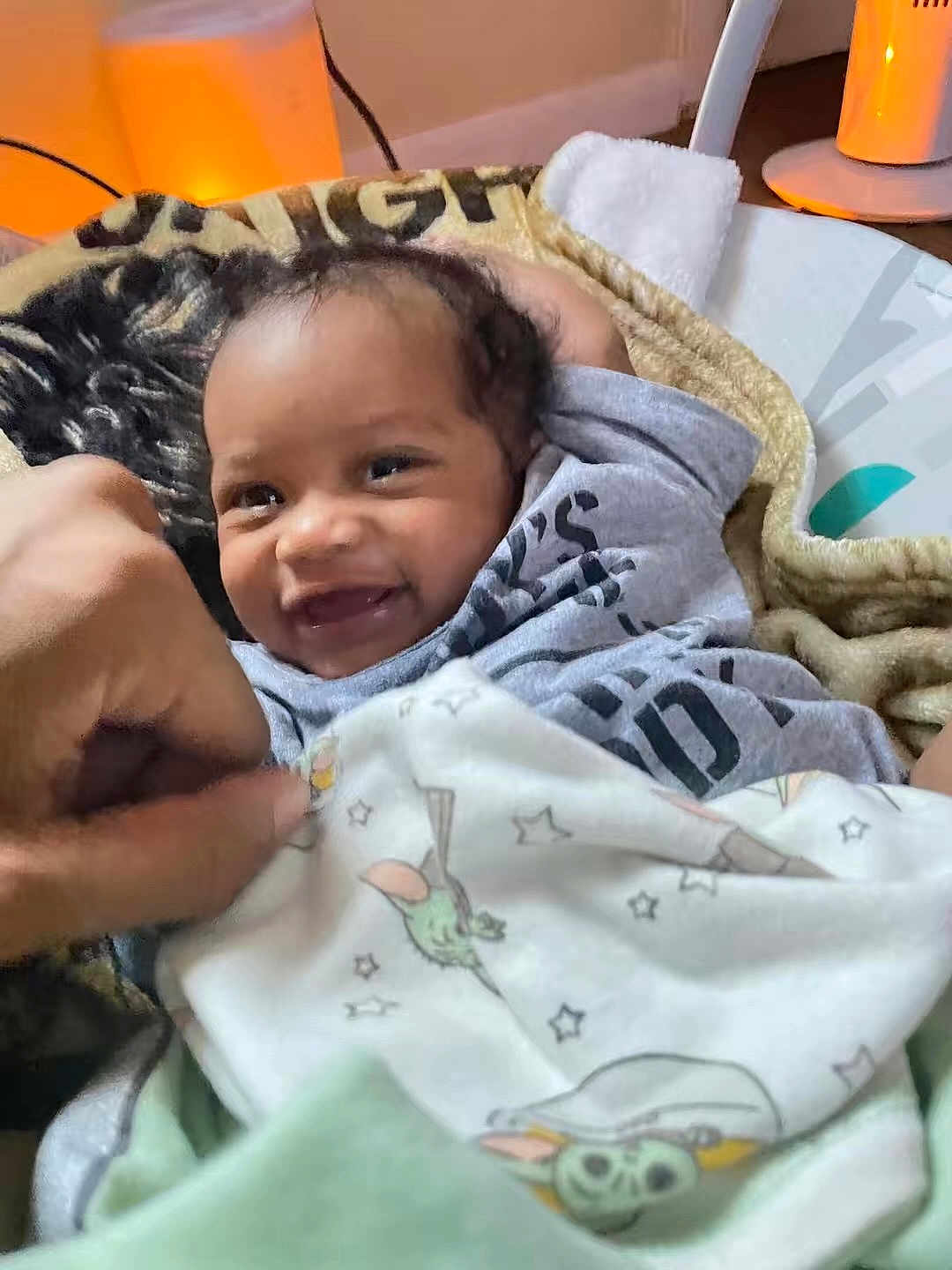 Nyzaiah is registered to the contest to win money with this photo: baby, smiling, blanket, hand, indoor, cozy, infant, clothing, face, person, cute, warm_light, toddler, expression, comfort, furniture, soft, portrait, happy, playful