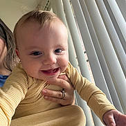 Connor is registered to the contest to win money with this photo: baby, child, infant, smiling, happy, portrait, close_up, indoor, window, blinds, sunlight, yellow_clothing, striped_outfit, adult_hand, caregiver, sitting, playful, face, home, photography