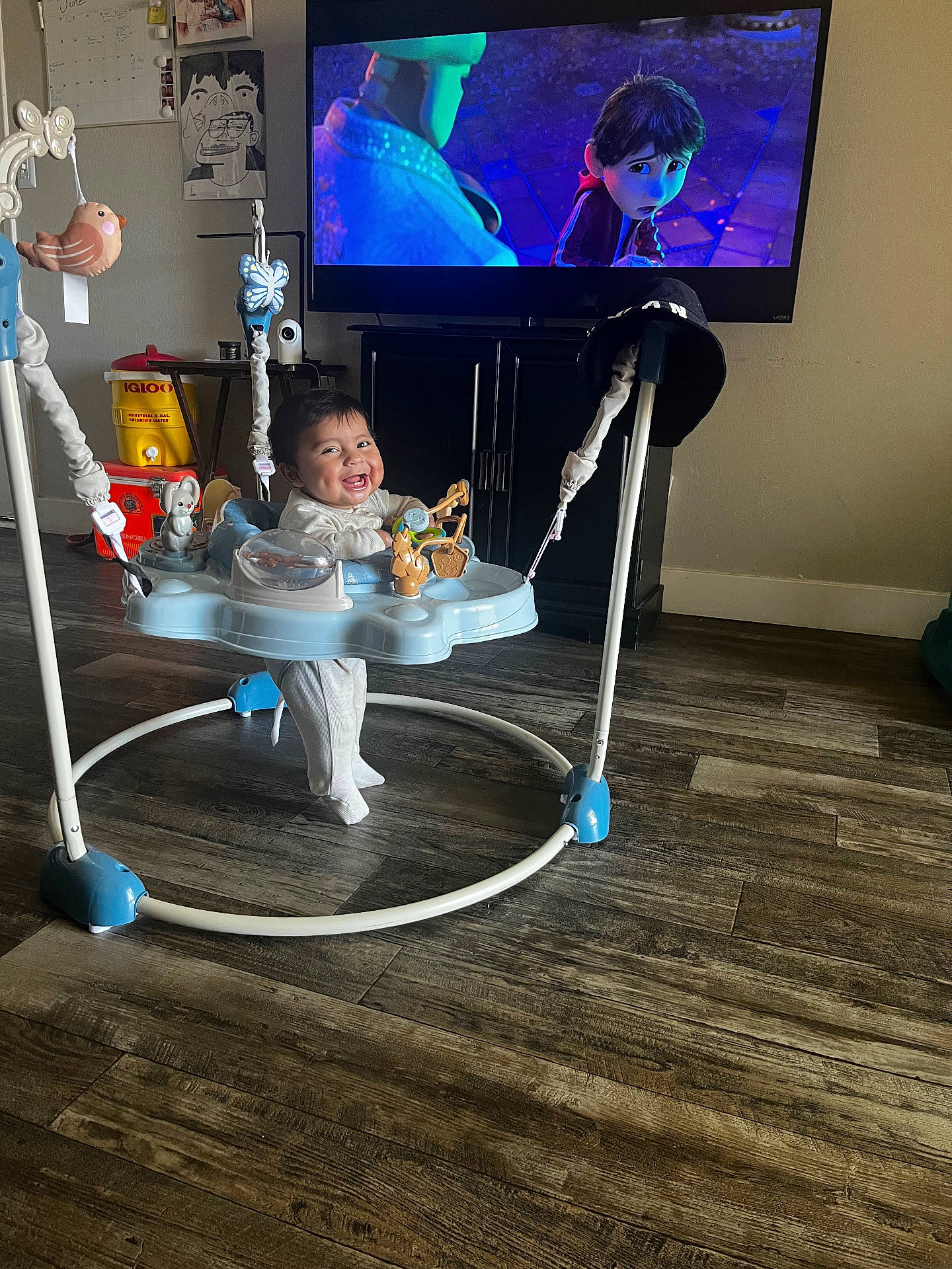 Valentino is registered to the contest to win money with this photo: animated_movie, baby, baby_jumper, cabinet, child, clothing, entertainment, flooring, furniture, happy, hat, home, indoor, living_room, person, screen, smiling, television, toys, wood_floor