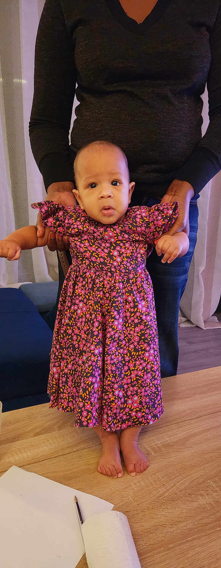 Fatima participe au concours pour gagner de l'argent avec cette photo : baby, child, person, adult_hand, floral_dress, standing, wooden_table, indoor, curtains, couch, footwearless, supporting_hands, long_sleeve_shirt, jeans, floor, furniture, portrait, expression, looking_at_camera, home