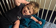 Elijah joined the competition — help win amazing prizes! toddler, child, crib, blanket, bed, blue, white, zigzag_pattern, smiling, happy, hair, clothing, shorts, shirt, indoor, person, foot, hand, face, playful