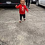 Kiara is registered to the contest to win money with this photo: toddler, child, smiling, red_shirt, gingerbread_man, walking, concrete, driveway, car, black_car, white_car, person, adult, happy, outdoor, daylight, footwear, curly_hair, accessory, casual_clothing