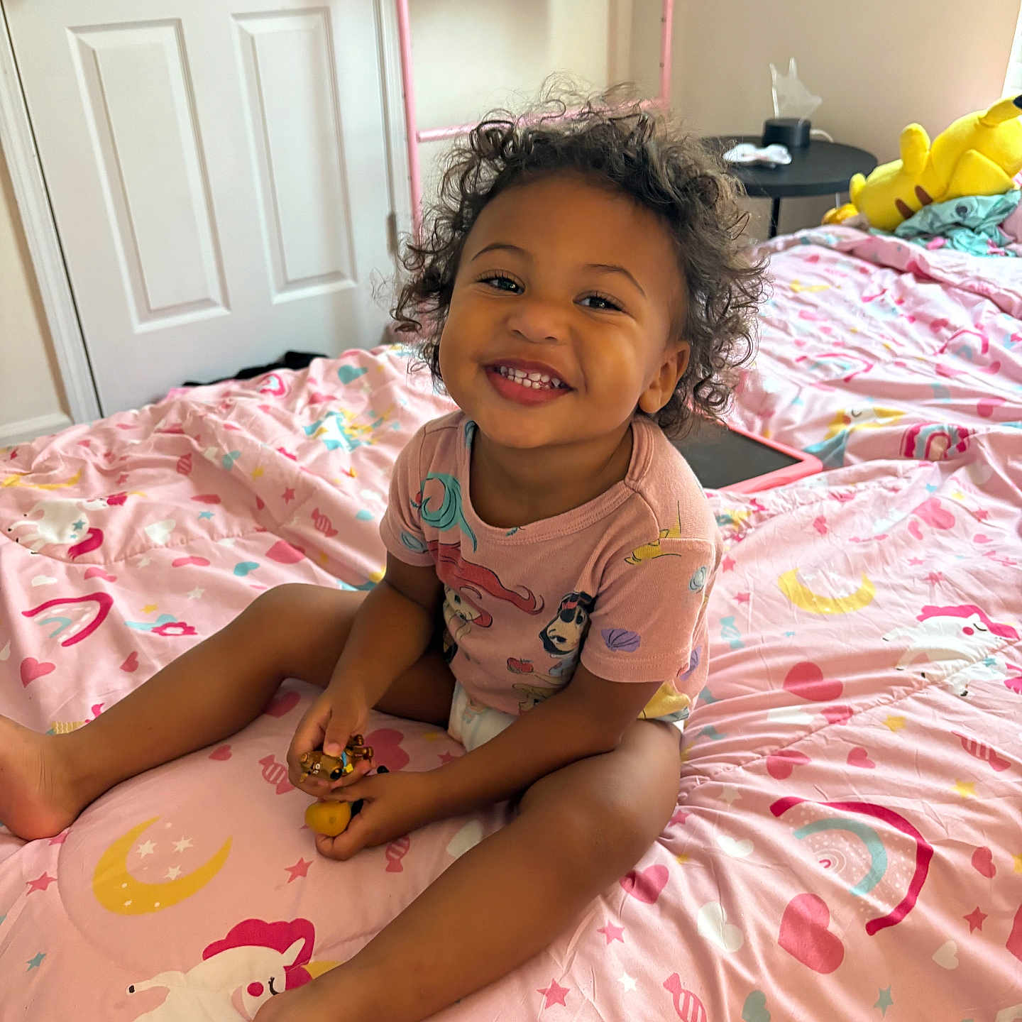 Kiara is registered to the contest to win money with this photo: bed, bedroom, child, clothing, curly_hair, door, furniture, happy, hearts, indoor, person, pink_bedspread, plush_toy, rainbows, sitting, smiling, toddler, towel, toy, unicorn