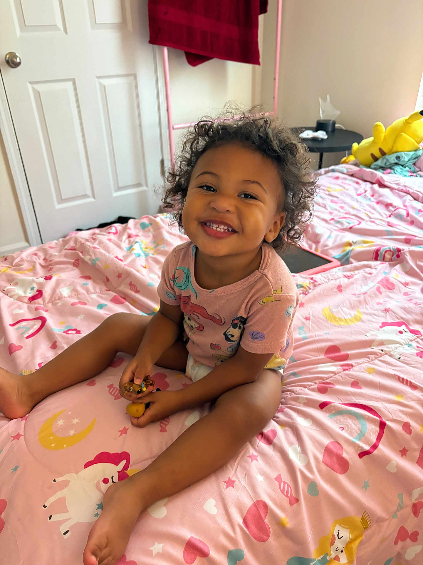 Kiara is registered to the contest to win money with this photo: toddler, child, smiling, curly_hair, bedroom, bed, pink_bedspread, unicorn, hearts, rainbows, toy, sitting, happy, clothing, indoor, furniture, door, towel, plush_toy, person
