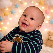 Titan is registered to the contest to win money with this photo: baby, background_lights, blurred_background, child, cozy, cute, face, football_patch, green_stripes, happy, indoor, infant, navy_blue, plush_toy, portrait, smiling, soft_lighting, star_decorations, striped_clothing, teddy_bear