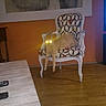 cat, chair, patterned_furniture, wooden_floor, indoor, living_room, glowing_eyes, furniture, pet, animal, orange_wall, table, remote_control, artwork, legs_dangling, cozy, domestic, home, relaxed, night