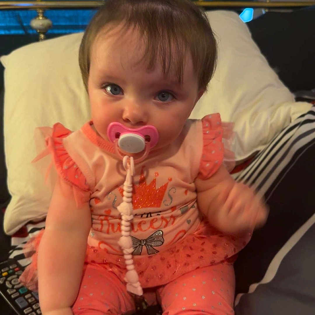 Lilyonna joined the competition — help win amazing prizes! baby, bed, child, clothing, curious, cute, hair_tie, indoor, infant, leggings, pacifier, person, pillow, pink_bow, plush, princess_outfit, remote_control, sitting, toddler, white_shoes