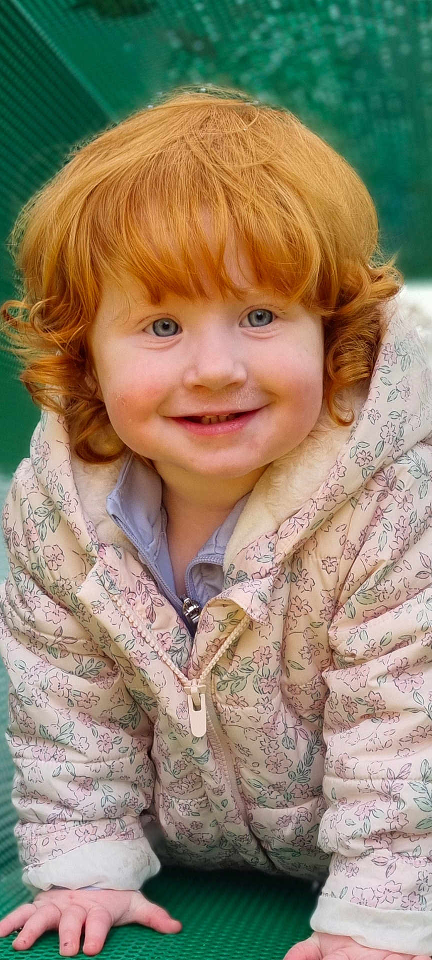 Rosie is registered to the contest to win money with this photo: toddler, child, red_hair, curly_hair, smiling, blue_eyes, jacket, floral_pattern, crawling, hands, outdoor, green_background, cute, happy, portrait, face, winter_clothing, innocence, rosy_cheeks, person