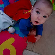 Valentin participe au concours pour gagner de l'argent avec cette photo : baby, child, blue_clothing, red_cape, toy, colorful_mat, playmat, floor, indoor, curious, wide_eyes, open_mouth, infant, puzzle_mat, soft_toy, necklace, hand, lying_down, superhero_costume, playtime