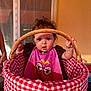 baby, basket, wicker, checkered_fabric, pink_bib, curious, indoor, child, holding, handle, wide_eyes, expression, hair, sunlight, shadow, mouth_open, seated, person, cute, portrait