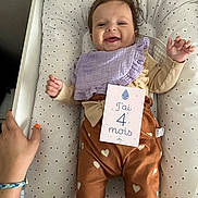 Lisa participe au concours pour gagner de l'argent avec cette photo : baby, infant, smiling, lying_down, cushion, clothing, bib, hand, milestone_card, heart_pattern, bed, person, cute, happy, indoors, fabric, skin, face, child, four_months
