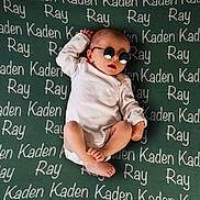 Kaden joined the competition — help win amazing prizes! baby, child, infant, sunglasses, white_onesie, blanket, text, names, lying_down, cute, cool, relaxed, foot, hand, head, skin, person, indoor, portrait, fashion