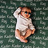 baby, child, infant, sunglasses, white_onesie, blanket, text, names, lying_down, cute, cool, relaxed, foot, hand, head, skin, person, indoor, portrait, fashion