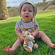 Timéo participe au concours pour gagner de l'argent avec cette photo : baby, child, grass, outdoor, plush_toy, tiger_toy, hat, sun_hat, greenery, nature, cute, sitting, infant, toy, person, summer, daylight, expression, face, legs