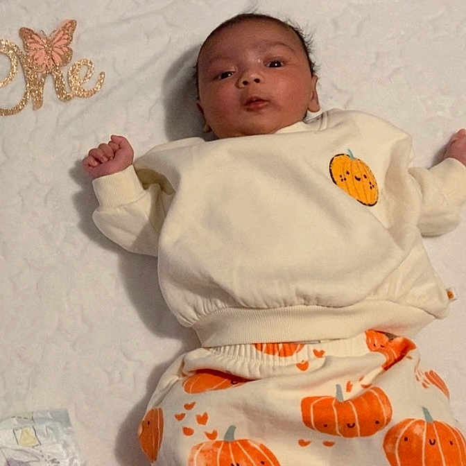 Gursidak Kaur is registered to the contest to win money with this photo: baby, blanket, celebration, clothing, cute, decor, expression, face, glitter, indoor, infant, lying_down, milestone, newborn, one, pants, person, pumpkin, soft_texture, sweatshirt