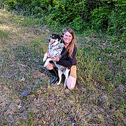 Tink is registered to the contest to win money with this photo: woman, dog, outdoor, grass, nature, greenery, smiling, kneeling, happy, pet, animal, casual_clothing, boots, trees, sunlight, spring, friendship, companionship, field, daytime